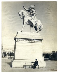 大きな台座の上に馬に座っているアメリカンインディアンの像を示す垂直のセピア色の写真。台座のベースのベンチに座っている黒いスーツと帽子の男 作： Unknown